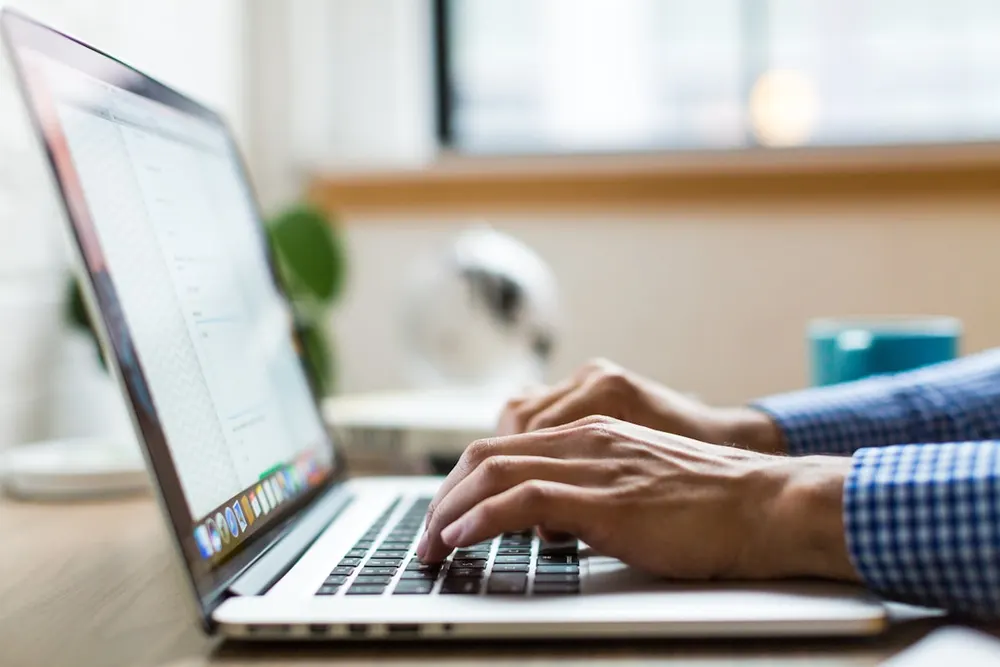 Hands typing on a laptop at a desk in an open office setting